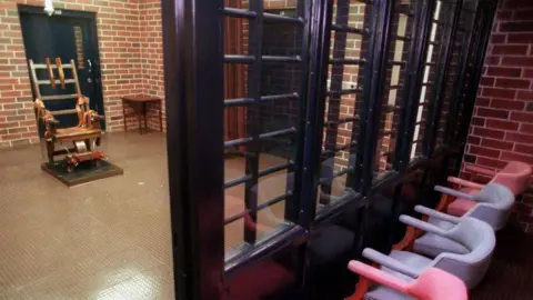 Getty Images Four chairs are positioned behind a black wall with lots of windows. The viewing area looks out onto a chamber with brick walls and an electric chair in the middle of the room.